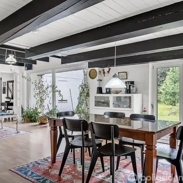 Spisebord med seks sorte stole står i et lyst rum med bjælkelofter og store vinduer, der åbner ud mod en grøn have. Tæpper og planter fuldender en hyggelig atmosfære.