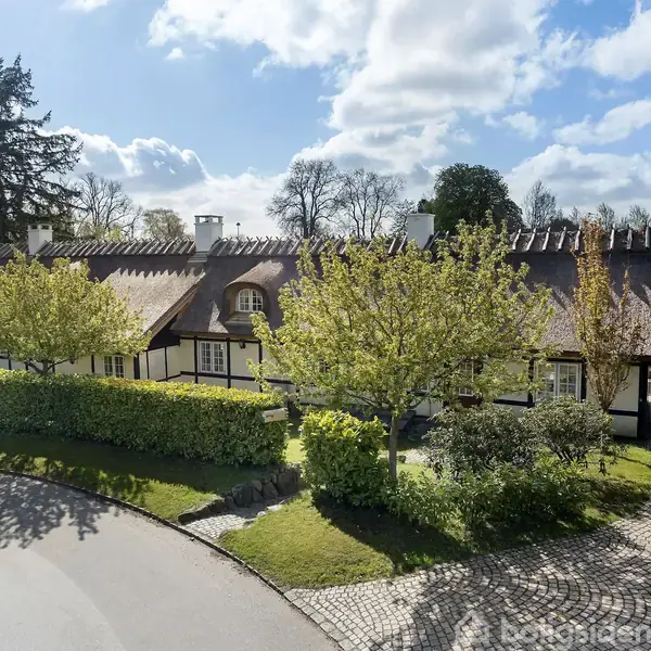 Et stråtækt hus med hvide vægge og sorte bindingsværk står omgivet af frodig have og træer. Det ligger ved en stille brolagt vej under en blå, skyet himmel.