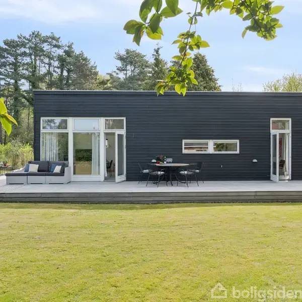 En moderne sort træbungalow står stille mod en baggrund af frodige træer. Foran huset er der en terrasse med en sofa og et bord, omgivet af en velplejet græsplæne.