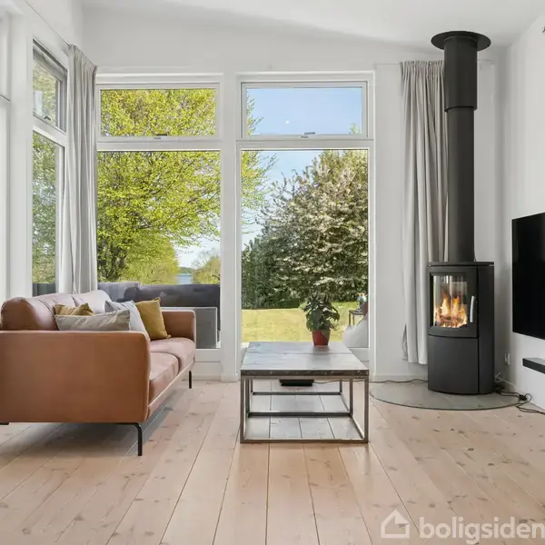 En stue med en brun sofa står roligt, mens en pejs brænder i hjørnet. Store vinduer afslører en grøn have. Der er også et fladskærms-tv på væggen.