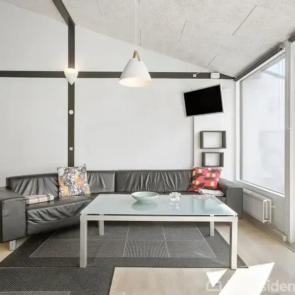 En sort lædersofa med farverige puder står i en moderne stue. En glasbord foran sofaen har en skål og en vase. Et fladskærms-TV hænger på væggen over hylder. Store vinduer giver lysindgang.