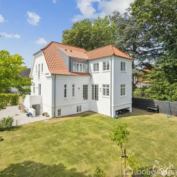 Hvid villa med rødt tegltag står stille på en velplejet græsplæne, omgivet af frodige træer og en sort plankevæg i en solrig have.