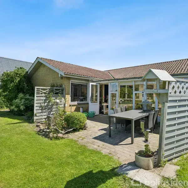 En gulstensvilla med rødt tegltag har en terrasse med havemøbler. Omgivet af grønne buske og græsplæne under en klar blå himmel, skaber det en hyggelig haveatmosfære.
