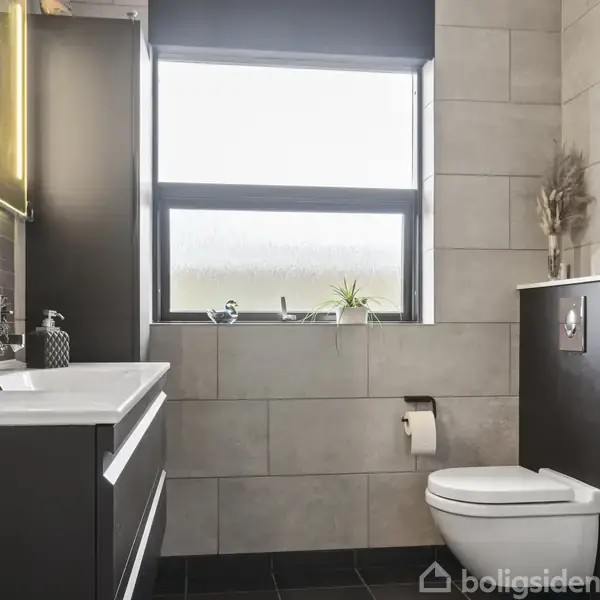 Et moderne enfamiliehus med fladt tag og flere vinduer står midt i en velplejet have med hække. I baggrunden ses en sø og flere træer under en klar himmel.