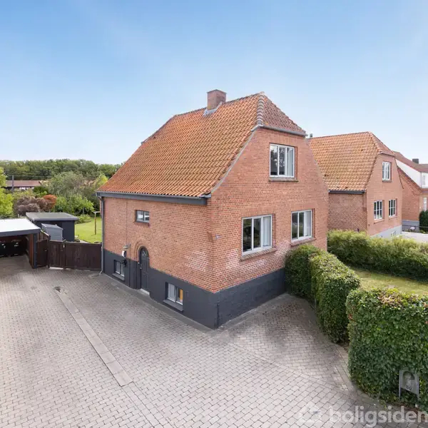 Rødt murstenshus står stille på en flisebelagt indkørsel omgivet af grønne hække i et villakvarter. Himlen er blå og klar.