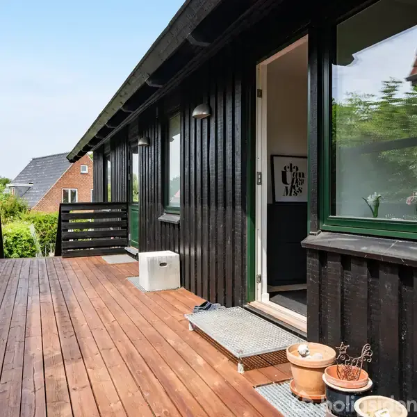 Sortmalet træhus med terrasse og åbent dørparti fører ind til huset. Der står potteplanter langs kanten. Omgivelserne har grønne buske og nabobygninger i baggrunden.