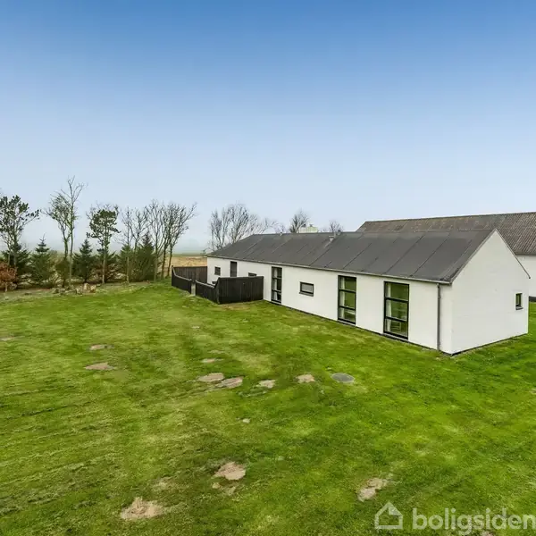 Hvidt enfamiliehus med mørkt tag står på stor, åben græsplæne. Træer og buske danner en grøn baggrund, mens himlen er klar og blå.