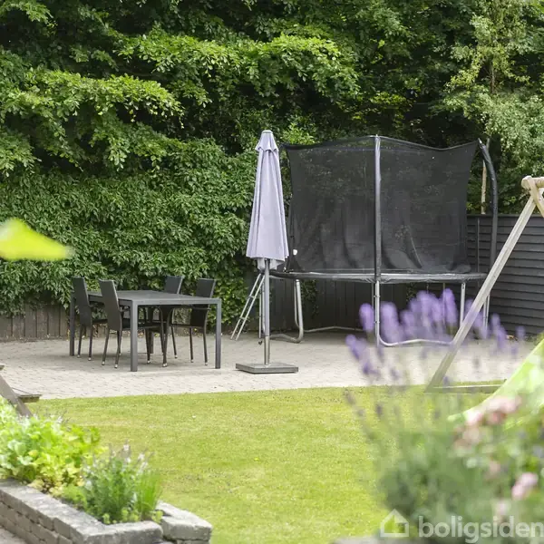 En trampolin står i en have ved siden af et bord- og stolesæt, omgivet af grønne planter og en legeplads med rutsjebane. En solparasol er placeret ved bordet.