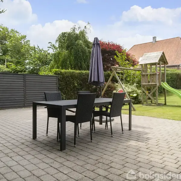 Et sort havebord med fire stole står på en flisebelagt terrasse. Ved siden af er en trampolin og en legeplads med rutsjebane. Omkring er der grøn beplantning og huse i baggrunden.