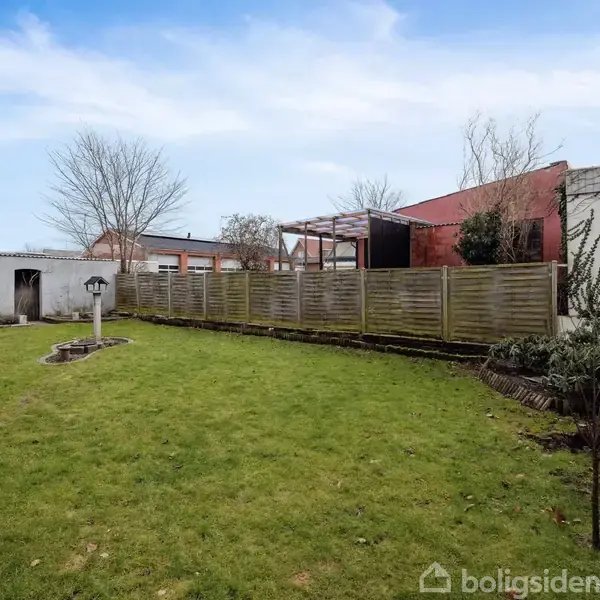 Græsplæne med en enkelt lanterne, omgivet af en plankevæg og mure i en baggård. Himmel er overskyet med bygninger i baggrunden og et par spredte træer.