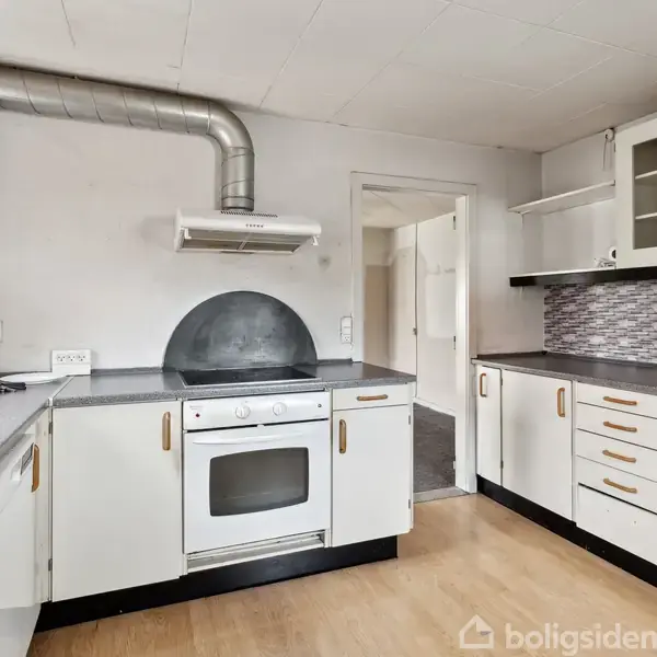 Komfur med integreret ovn står i et lyst køkken. Køkkenet har hvide skabe, sorte bordplader, opvaskemaskine, emhætte og mosaikflise backsplash, med en døråbning til en anden del af huset.