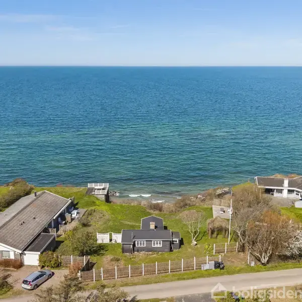 Feriehus ved kysten, omringet af grønne træer og buske, ligger ved en vej. Huset har havudsigt med blåt vand i baggrunden.