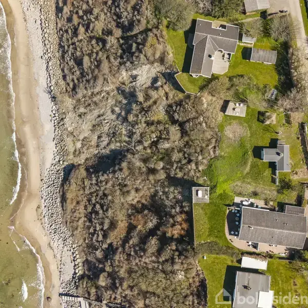 Kystlinje med grønklæde huse ved siden af en strand. Huse er omgivet af veje og vegetation. Havet er til venstre med bølger, der rammer den stenede strand.