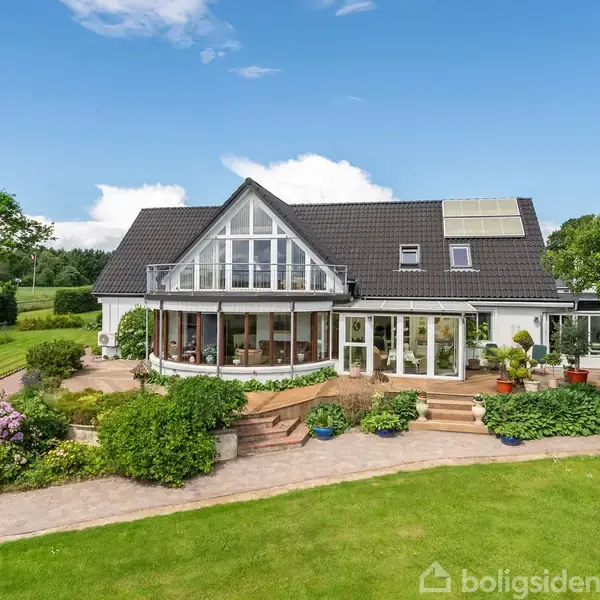 Hus med stort glasparti og gråt tag, omgivet af velplejet have med blomsterbede og græsplæne. Solpaneler på taget, trapper fører ned til stenlagt terrasse. Skyfri blå himmel.