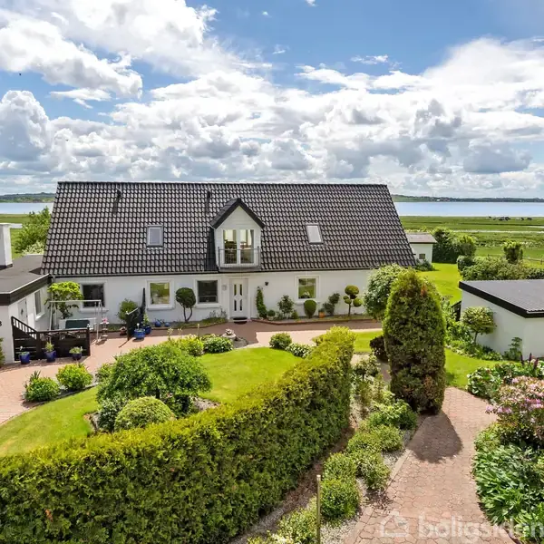 Et stort, hvidt hus med sort tegltag står omkranset af en velholdt have. I baggrunden ses en vidstrakt udsigt over marker og en sø under en let overskyet himmel.