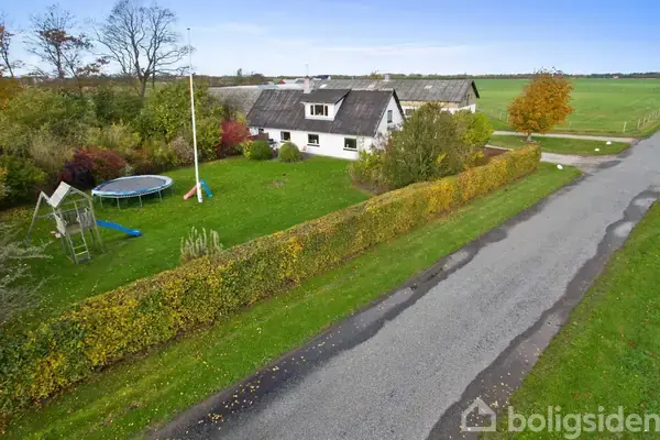 Hus omgivet af grøn have med trampolin og gynge. Handling sker nær en landevej, omringet af buske og træer. Landskabet består af åbne marker i baggrunden.