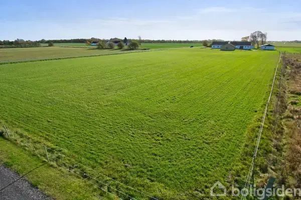 Græsmark strækker sig bredt med en række bygninger i baggrunden, omgivet af marker og træer under en klar blå himmel. Et hegn løber langs kanten af marken.