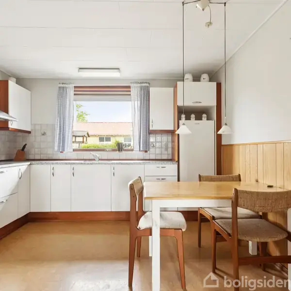 Køkken med hvide skabe og stål-emhætte. Et bord med to stole står foran et vindue med udsigt til en have. Trævægspaneler skaber et klassisk udseende.