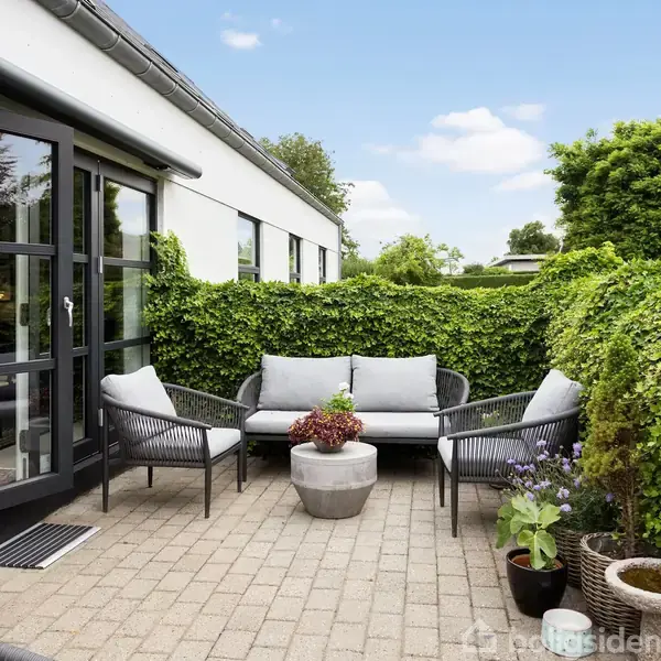 Terrassemøbler på flisebelagt patio med sofa og stole arrangeret omkring et rundt bord. Huset har sorte vinduesrammer. Baggrunden er grønne buske og planter under en blå himmel.