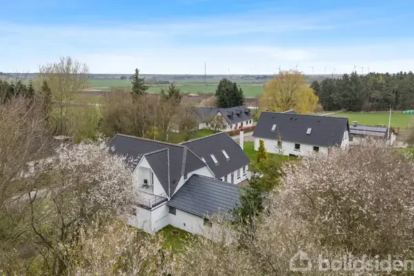 En gruppe hvide huse med sorte tage ligger blandt træer i en landlig grøn landskab. Huse er omgivet af marker og skov, under en klar blå himmel.