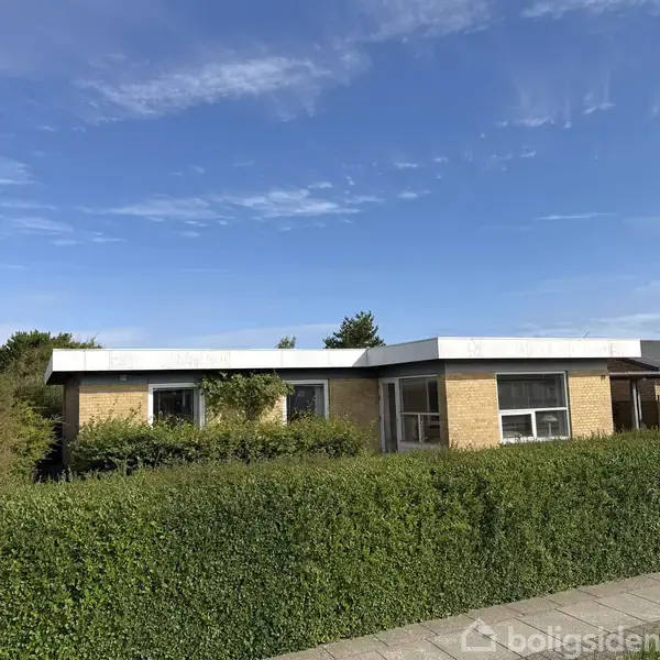 En enkeltstående, fladtaget murstenshus står bag en veltrimmet grøn hæk. Blå himmel med spredte skyer ses i baggrunden.