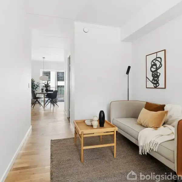 En sofa med tæppe og puder står i en lys stue. Et lille bord med skulpturer er foran sofaen. I baggrunden ses et spisebord med stole.