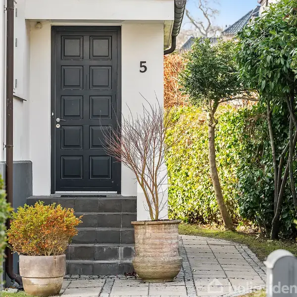 En sort dør med nummer 5 er omgivet af krukker med planter. Døren fører til et hus, og en flisebelagt sti er flankeret af buske og hække i haven.