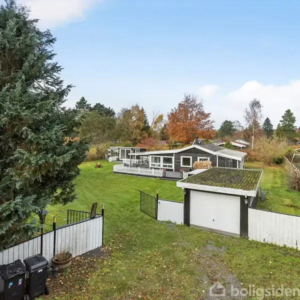 Hus omgivet af grønne planter og træer står stille i en have med hvidt hegn og en mindre garage. Landskabet er præget af efterårsløv og spredte træer i baggrunden.