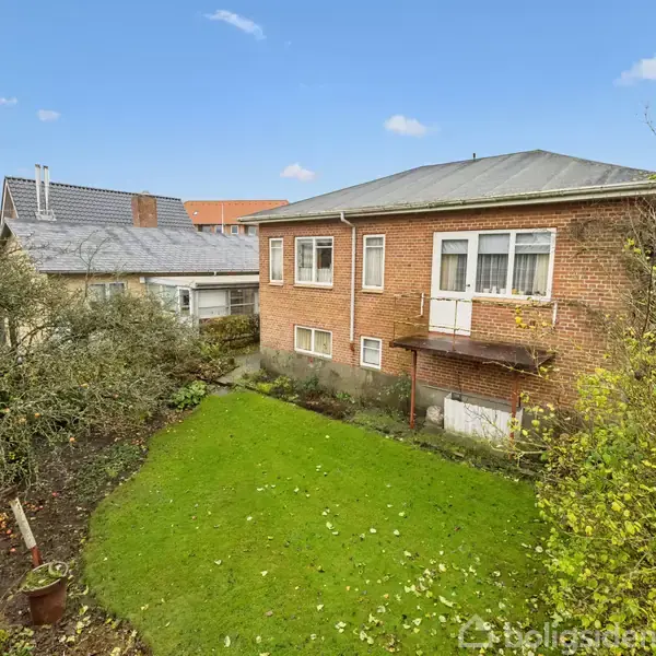 En murstensbygning med en lille balkon står over en velplejet græsplæne i en have. Baggrunden viser andre hustage under en klar blå himmel med få skyer.