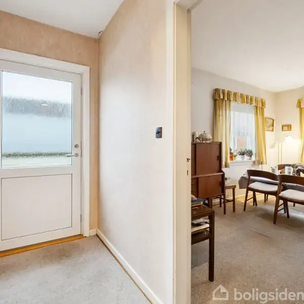 En gang med en glasdør til venstre fører ind til en stue med møbler og gardiner. Stuen har gulv-til-loft-vinduer og planter, hvilket skaber en hjemlig atmosfære.