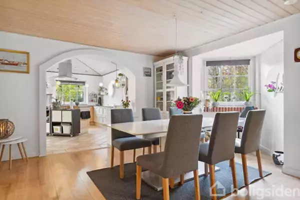 Et spisebord med seks grå stole står i et lyst rum med trægulv. I baggrunden ses et køkken med hvidt interiør. Store vinduer giver udsigt til træer og planter.