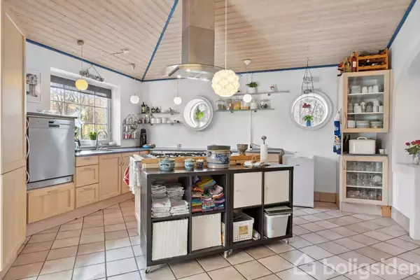 Et åbent køkken med en central ø og hylder, indeholder moderne apparater. Der er dekorative lamper og et vindue med udsigt til en have.