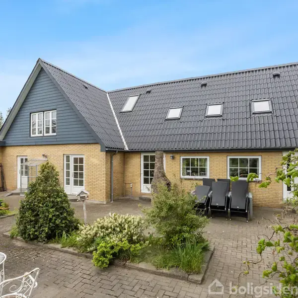 Et moderne hus med skråt tag og ovenlysvinduer står i en frodig have med fliser, buske, og havemøbler. Himlen er klar og blå i baggrunden.
