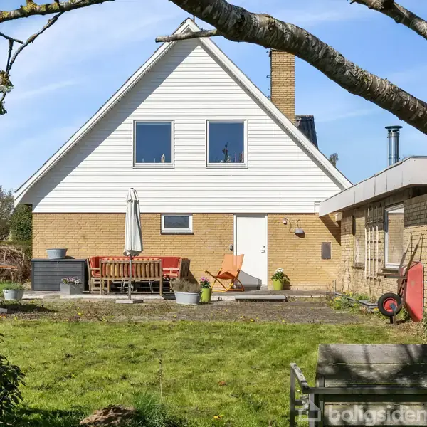 Hus med gavl tag, to store vinduer, og terrasse på bagsiden. Røde stole og bord er placeret på terrassen. Græsplæne omgiver huset og en stor trægren hænger i forgrunden.