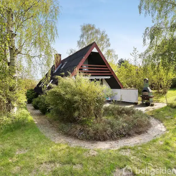 A-formet hus med sort tag står stille i frodig have med træer og buske. Stien fører til terrasse med udendørs grillområde.