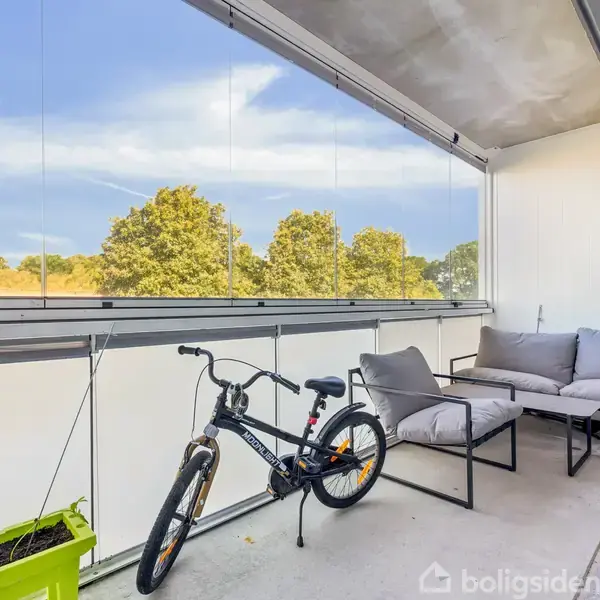 Cykel står stille på altan med glasafskærmning; grå sofaer står til højre, planter i en grøn kasse til venstre. I baggrunden ses trætoppene og en blå himmel.