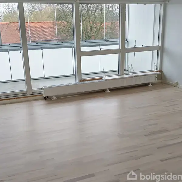 Et tomt rum med lyst trægulv, store vinduer, og en radiator under vinduerne. Udsigt til røde tage og træer udenfor.