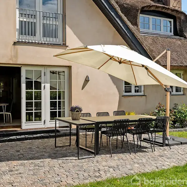Terrasse med spisebord og otte stole under en stor parasol. Hus med stråtag og terrasse i brosten som kontekst. Græsplæne og buske omgiver området.