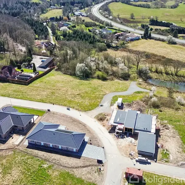 Bygninger under konstruktion ved en snoet vej omgivet af marker og skov i naturskønt område. Lille sø og spredte boliger i baggrunden.