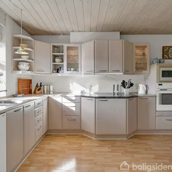 Et lyst køkken med hvide skabe og naturligt lys, der strømmer ind gennem vinduet. Køkkenet rummer en ovn, kogeplader og diverse køkkenredskaber, omgivet af en trælaminatgulv.