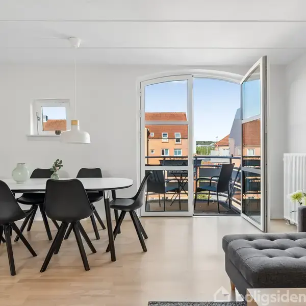 En moderne spisestue med sort spisebord og seks sorte stole. En dør står åben til en balkon med udsigt over nærliggende bygninger. Sofa til højre er dekoreret med farvet pude.