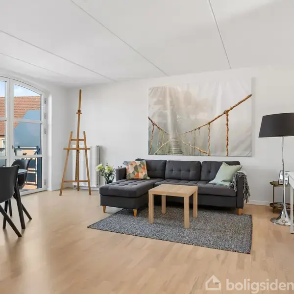 Stue med sort sofa og sofabord placeret på tæppe, vægmaleri af hængebro ovenfor. Spisebord med fire sorte stole til venstre, vindue bagved viser hustage.