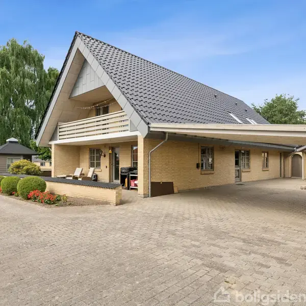 Et gulstenshus med mørkt tag står i en velplejet have. Huset har en overdækket terrasse med liggestole. En stor indkørsel fører op til carporten. Solrig dag.