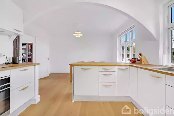 Et lyst køkken med hvide skabe og træbordplader står i centrum. Det er roligt med store vinduer, der lukker naturligt lys ind. Gennemsigtig frugtskål og knivblok i hjørnet.