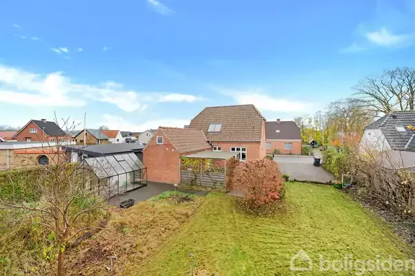 Et rødt murstenshus står i en have med en græsplæne og nogle buske. Til venstre ses et drivhus. Nabohuse omkranser området under en blå himmel.