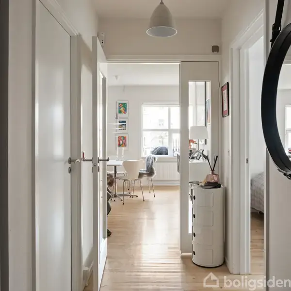 En lys gang med trægulv leder ind til en spisestue. Gangen har hvide døre, kommoder med dekorationer, og et rundt spejl til højre.