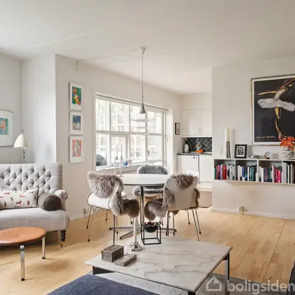 Et lyst stueområde med moderne indretning. En grå lænestol ved siden af et lille bord. Et spisebord med pelsbeklædte stole står centralt. Reoler med bøger og kunst på væggene.