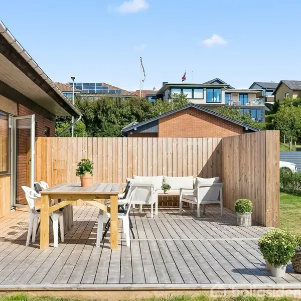 Terrasse med træbord og stole, dekoreret med planter, står ved et murstenshus. I baggrunden ses træplankeværk, græsplæne og flere farverige huse.