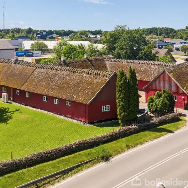 En stor rød lade med stråtag står ved en vej i et grønt landskab. Der er nogle høje træer ved siden af bygningen.