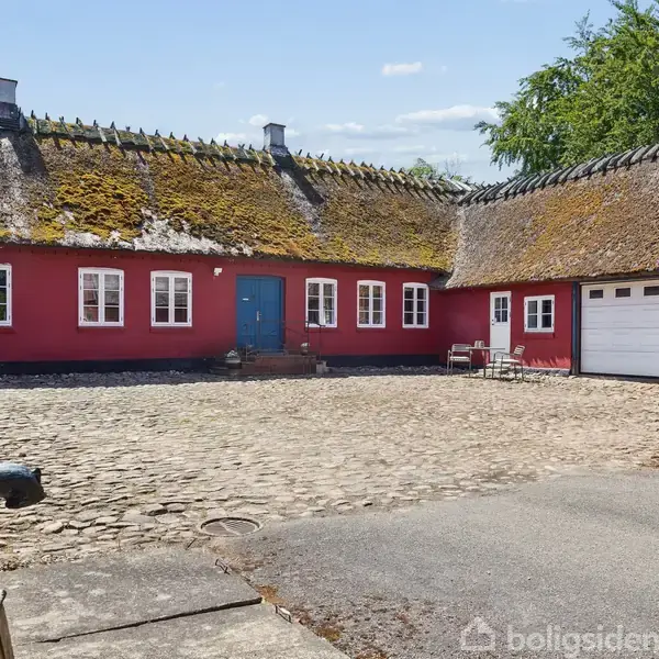En rød bindingsværksgård med stråtag omkranser en brostensbelagt gårdsplads. En sort vandpumpe står i forgrunden. Baggrunden viser grønne træer under en klar blå himmel.